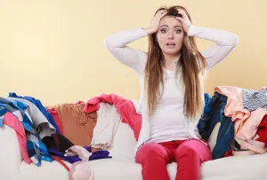 Woman frustrated holding her head sitting on couch surrounded by clothes